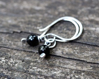 May include: A pair of dangle earrings featuring small, shiny black beads suspended from silver-colored hooks. The earrings have a simple, minimalist design, with the beads appearing to be round and smooth. The hooks are curved and have a classic fish hook style.