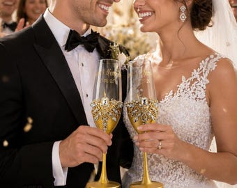 May include: A couple smiles, holding champagne flutes at a wedding. The gold-stemmed flutes are adorned with ornate gold and crystal embellishments. The bride wears a tiara and a white lace wedding dress, while the groom wears a black tuxedo.