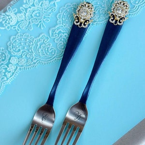 May include: Two blue forks with gold and rhinestone embellishments. The forks are on a light blue background with white lace trim. The forks are engraved with the letters "S" and "D".