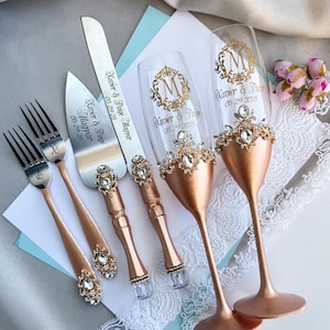 Könnte beinhalten: Ein Hochzeitstorten-Servierset und Champagnergläser. Das Set enthält ein Tortenmesser, einen Servierer und zwei Gabeln mit roségoldenen Griffen und Kristallakzenten. Die Champagnergläser sind roségoldfarben mit Kristallverzierungen.