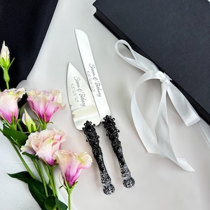 Puede incluir: Un juego de cuchillo y servidor para pastel de bodas con mangos negros ornamentados y texto grabado. Las hojas son plateadas y los mangos tienen un diseño decorativo detallado. El juego está junto a un ramo de flores rosas y blancas.