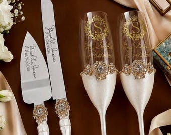 May include: A cake knife and server set with white handles and gold accents, paired with two champagne flutes featuring gold monogrammed S initials, floral embellishments, and white stems, all engraved with the names Yagid and Savene and 16/04/2026.