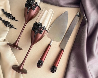 May include: Two burgundy champagne flutes with black lace and rhinestone embellishments, a burgundy cake knife with black rhinestone embellishments, and a silver cake server.