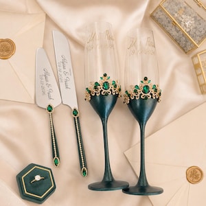 May include: Wedding set featuring two champagne flutes with emerald green and gold embellishments, a cake knife and server, and a ring in a green box. The flutes are inscribed with names and a date.