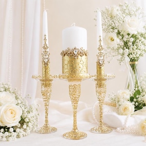 May include: Three white candles set in ornate gold-toned glass holders. The two outer taper candles feature gold filigree accents with clear crystals. The central pillar candle has a gold base with swirling patterns and a crystal-encrusted band.