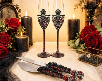 May include: Two tall champagne flutes and a matching cake serving set featuring intricate black and red filigree detailing. The glasses have slender stems and ornate embellishments, while the knife and server handles mirror the same pattern.