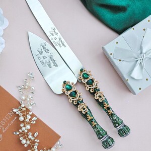 May include: A silver cake knife and server set featuring ornate handles with dark green and gold accents, embellished with clear crystals. The blades are engraved with the text Anette Santiago Mis 15 Años 3/13/2027 in a script font.