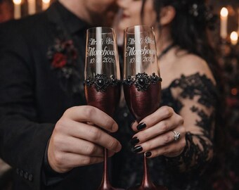 May include: Two champagne flutes with burgundy stems and bowls. The bowls are decorated with black filigree and the text "Mr & Mrs Morehouse 10 2026". The flutes are held in front of a blurred background of a couple.
