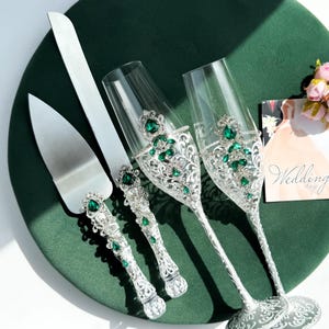 May include: A wedding cake knife and server set with two champagne flutes. The silver handles are adorned with emerald green jewels and clear rhinestones. The flutes have a decorative silver design with emerald green jewels. A wedding day card is in the background.