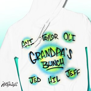 May include: White hoodie featuring airbrushed text and graphics. The design includes the words "GRANDPA'S BUNCH" in a blue and yellow circle, surrounded by names such as "ANT", "TAYLOR", "OLI", "JED", "HIL", and "JEFF".