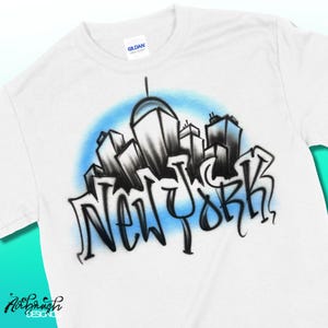May include: White t-shirt featuring a stylized black and white cityscape design with the word "NEW YORK" in graffiti-style lettering. The design is set against a light blue background. The shirt has a crew neck and short sleeves.