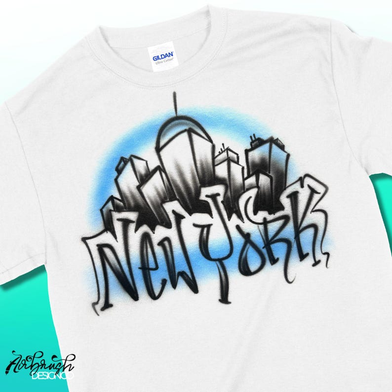 May include: White t-shirt featuring a stylized black and white cityscape design with the word "NEW YORK" in graffiti-style lettering. The design is set against a light blue background. The shirt has a crew neck and short sleeves.