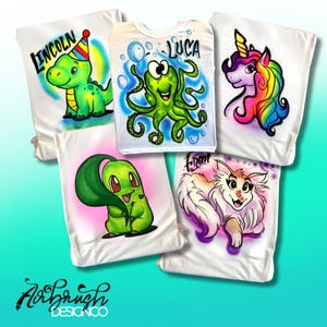 Könnte beinhalten: Weiße T-Shirts mit Airbrush-Cartoon-Designs. Eines zeigt einen grünen Dinosaurier mit dem Namen "LINCOLN", ein anderes einen Oktopus mit "LUCA", ein Einhorn, eine grüne Kreatur und einen flauschigen Hund mit "Fran". Das Logo "Airbrush Design Co" befindet sich unten.
