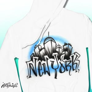 May include: White hooded sweatshirt featuring a black and gray skyline design with the text "New York" in a graffiti style. The design is set against a light blue background.