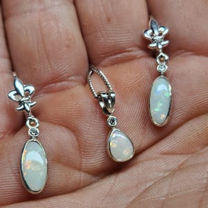 Puede incluir: Un conjunto de joyas con un collar con colgante de plata y pendientes a juego. Los pendientes y el colgante tienen un diseño de flor de lis en la parte superior, con una pequeña gema redonda y una piedra de ópalo ovalada. Las piedras de ópalo tienen un brillo nacarado.