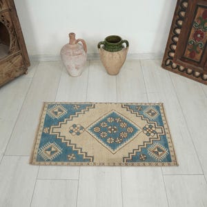 May include: A rectangular rug with a central diamond pattern in beige and blue. The rug features geometric designs and a border in a lighter beige colour. The rug is placed on a light-coloured wooden floor.