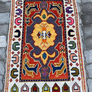 Pode incluir: Um tapete vintage colorido com um padrão geométrico em tons de vermelho, laranja, amarelo, azul, verde e roxo. O tapete tem uma borda branca com franjas.