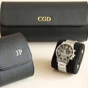 Puede incluir: Estuches negros para relojes y un reloj de pulsera plateado. El reloj tiene una esfera negra con detalles plateados y una correa plateada. Los estuches son negros con iniciales doradas. Un estuche es cilíndrico, el otro cuadrado.