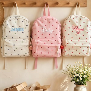 Può includere: Tre zaini appesi a un appendiabiti in legno. Quello a sinistra è color crema con la scritta blu James. Quello centrale è rosa con la scritta bianca Daisy. Quello a destra è color crema con la scritta rossa Naomi. Tutti hanno motivi a fragole.