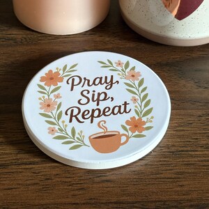 May include: A white coaster with the text "Pray, Sip, Repeat" surrounded by a floral wreath and a coffee cup illustration. A peach-colored candle and a speckled mug with a heart design are also visible.