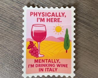 May include: A rectangular sticker with a pink border and the text "PHYSICALLY, I'M HERE. MENTALLY, I'M DRINKING WINE IN ITALY." The sticker features a wine glass, grapes, a sun, and a landscape with yellow fields and a green tree.