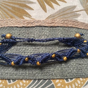 Può includere: Un braccialetto in macramè blu navy con perline dorate. Il braccialetto è realizzato con una tecnica di annodatura e ha un design semplice.