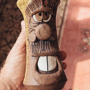 May include: A brown ceramic mug featuring a sculpted face with a prominent nose, textured eyebrows, a mustache, and two large white teeth visible in an open mouth. The exterior has a speckled tan finish on the sides and a dark brown clay texture.