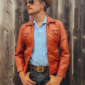May include: A rust-colored leather jacket with a zipper closure, worn over a light blue button-down shirt. The jacket has two chest pockets and a brown leather belt with a gold buckle. The person is wearing sunglasses and a hat.