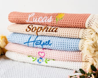 May include: A stack of waffle-weave towels in various colors, each embroidered with a name. The top towel is coral with the name "Lucas" and a yellow flower. Other towels are beige, blue, pink, and white.