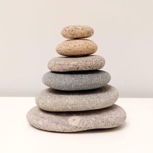 Può includere: Una pila di sei pietre lisce e arrotondate in varie tonalità di grigio, beige e marrone. Le pietre sono impilate l'una sull'altra, creando una torre. Lo sfondo è una superficie bianca.