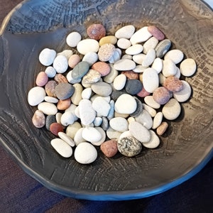 Può includere: Una ciotola nera piena di pietre lisce di colore grigio, bianco e marrone. Le pietre sono di diverse dimensioni e forme.