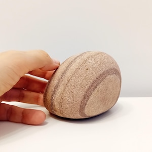 Puede incluir: Una piedra lisa de forma ovalada con una base de color marrón claro y bandas marrones más oscuras. Se aprecia la textura natural y el patrón único de la piedra. La piedra es sostenida por una mano. Un objeto decorativo natural.