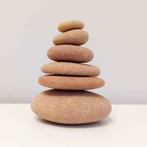 May include: A stack of six smooth, rounded stones in varying sizes, balanced atop one another. The stones range in color from light tan to reddish-brown, creating a natural, earthy aesthetic. The arrangement suggests balance and tranquility.