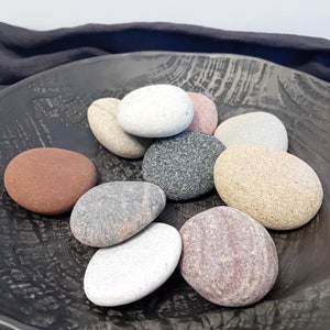 May include: A collection of smooth, round stones in various shades of gray, brown, and white, arranged in a black bowl. The stones are a natural, earthy element that can be used for decoration or relaxation.
