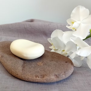 Puede incluir: Una pastilla de jabón blanca se encuentra sobre un jabonera de piedra marrón. Hay flores de orquídea blancas en el fondo.