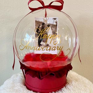 May include: A clear, round balloon with a red base and bow, containing a photo and red feathers. The photo features a couple, and the balloon has gold text that reads "Happy Anniversary."