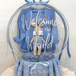 May include: A baby gift set in a clear balloon, featuring a blue sweater, a knitted bunny, and a blue blanket. The balloon has the text "Welcome to the World". The blanket has the text "Sweet Little Baby Marcel".