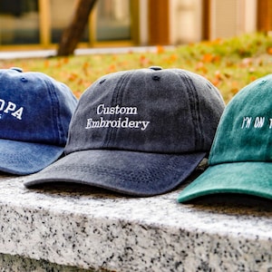 May include: Three baseball caps with embroidered text. The first is blue with "GRANDPA EST.2026", the second is dark grey with "Custom Embroidery", and the third is green with "I'M ON THE MARKET!".