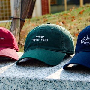 May include: Three baseball caps in a row, each with a different colour and text. The first is maroon with "I'm on the market!" in white. The second is green with "YOUR TEXT/LOGO" in white. The third is navy blue with "GRANDPA EST.2026" in white.