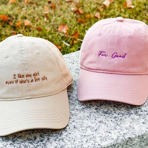 May include: Two baseball caps are shown, one beige and one pink. The beige cap has the embroidered text "I like my girl even if she's a bit silly." The pink cap has the embroidered text "For Good."