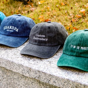 May include: Several baseball caps in shades of blue, grey, and green, each with embroidered text. The caps are arranged on a stone surface, with a backdrop of grass and autumn leaves. The text on the caps includes "awesome", "GRANDPA EST. 2026", "Custom Embroidery", and "I'M ON THE MARKET!".