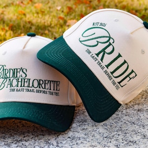 May include: Two baseball caps with cream-coloured crowns and dark green peaks. One cap says "Birdie's Bachelorette" in green script, and the other says "Bride". Both caps have the text "The Last Trail Before The Veil".