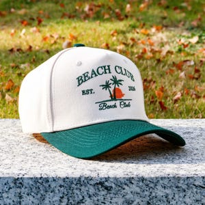 May include: A white and green baseball cap with the text "BEACH CLUB" embroidered on the front. The cap has a curved peak and a green button. The design includes palm trees and a sunset.