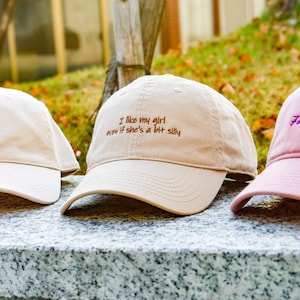 May include: Three baseball caps in a row. The first is beige with a bird graphic, the second is beige with the text "I like my girl even if she's a bit silly", and the third is pink with the text "For Good".