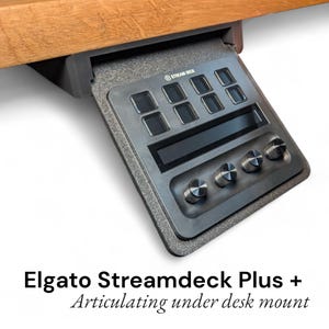 May include: A black Elgato Stream Deck Plus with an articulating under-desk mount. The device has multiple square buttons, a display screen, and four rotary knobs. The mount is attached to a wooden surface.