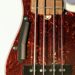 Könnte beinhalten: Nahaufnahme eines Basses mit einem Schildpatt-Pickguard und einem schwarzen Tonabnehmer. Die Saiten sind sichtbar und die Gitarre hat eine natürliche Holz-Oberfläche.
