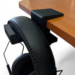 Könnte beinhalten: Schwarzer Kopfhörerständer mit einer Klemme, die an einem Schreibtisch oder Tisch befestigt wird. Der Ständer ist aus Kunststoff und hat eine schwarze Oberfläche. Der Ständer ist so konzipiert, dass er Kopfhörer sicher an Ort und Stelle hält.