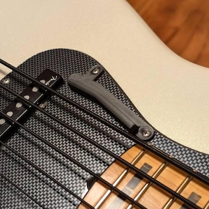 May include: Close-up of a black and white carbon fibre bass guitar with a black pickup and a black strap button. The guitar has a white body and a natural wood fretboard.