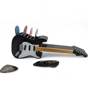 May include: A black electric guitar-shaped pick holder with a white pickguard. The holder has slots for multiple guitar picks. There are four picks in the holder, and three more picks are lying on a white surface.