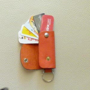 Puede incluir: Un llavero de cuero naranja brillante con un anillo de metal y una solapa que se abre para guardar varias tarjetas. Las tarjetas incluyen una tarjeta de cr&eacute;dito, una tarjeta de fidelizaci&oacute;n de la tienda y una tarjeta de seguro m&eacute;dico.
